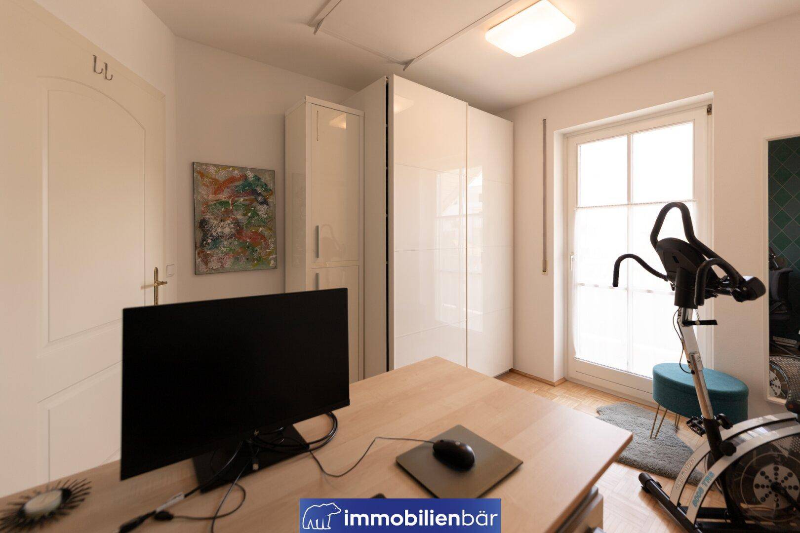 Helles Arbeitszimmer mit weißen Schränken, Schreibtisch und Sportgerät.