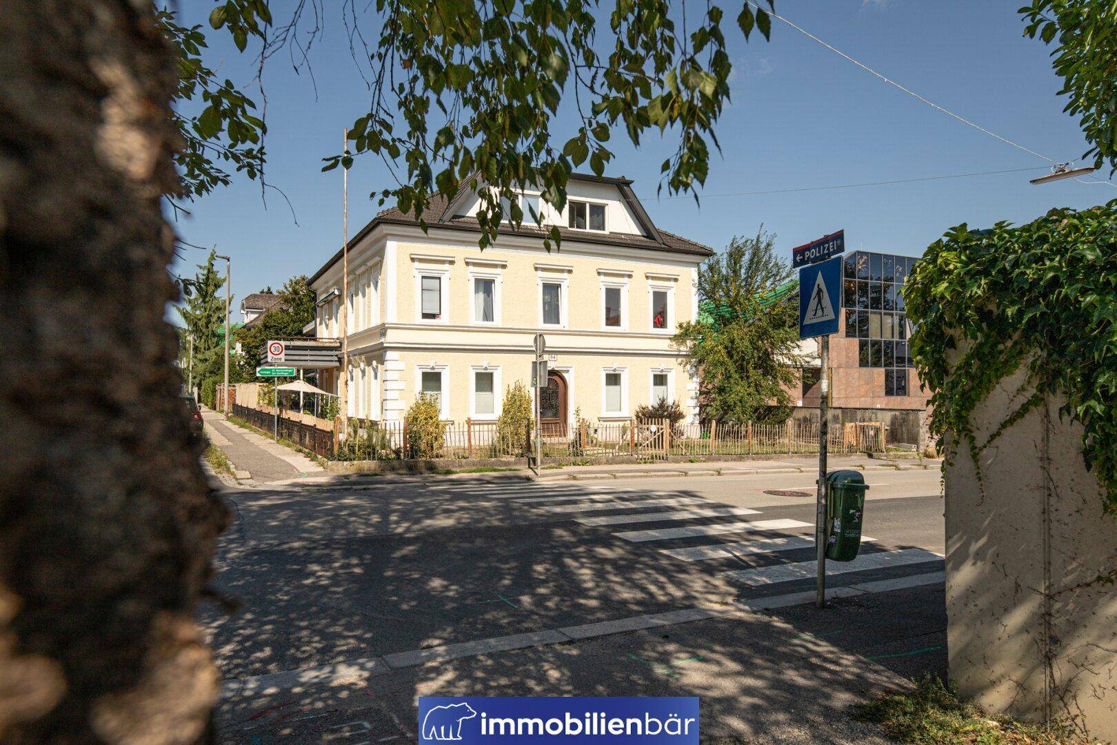Das historische Gebäude von der Straße aus gesehen, teilweise verdeckt von grünen Baumblättern.