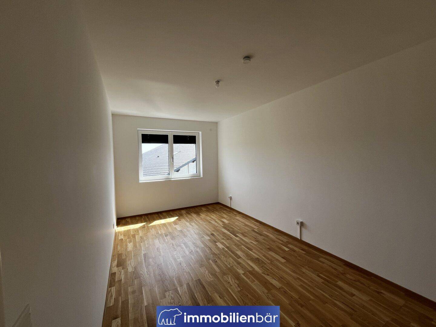 Helles Schlafzimmer mit Parkettboden und einem Fenster mit Blick ins Grüne.