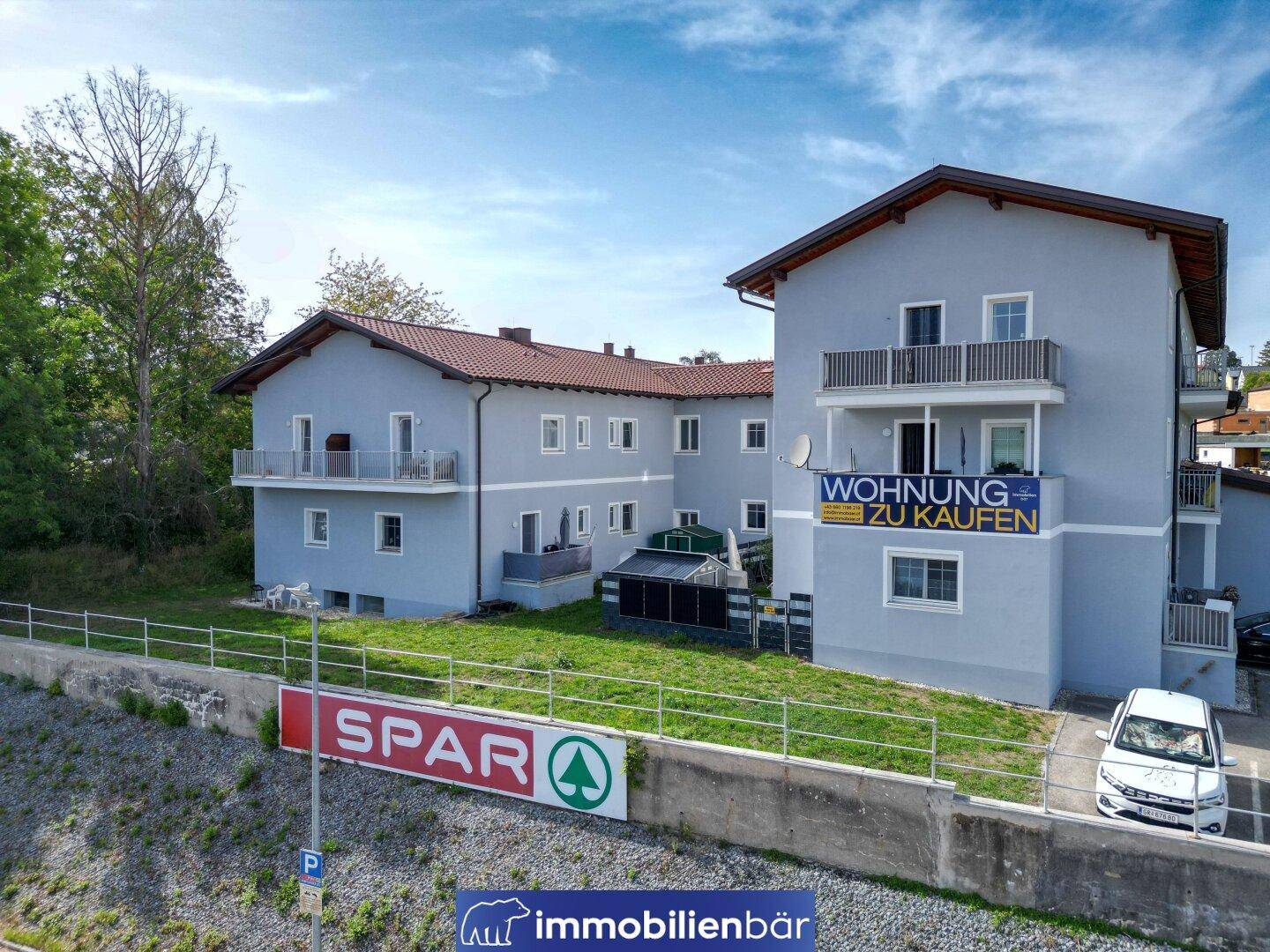 Panoramablick auf ein Wohngebäude mit hellgrauer Fassade, roten Dächern und Balkonen in ländlicher Umgebung.
