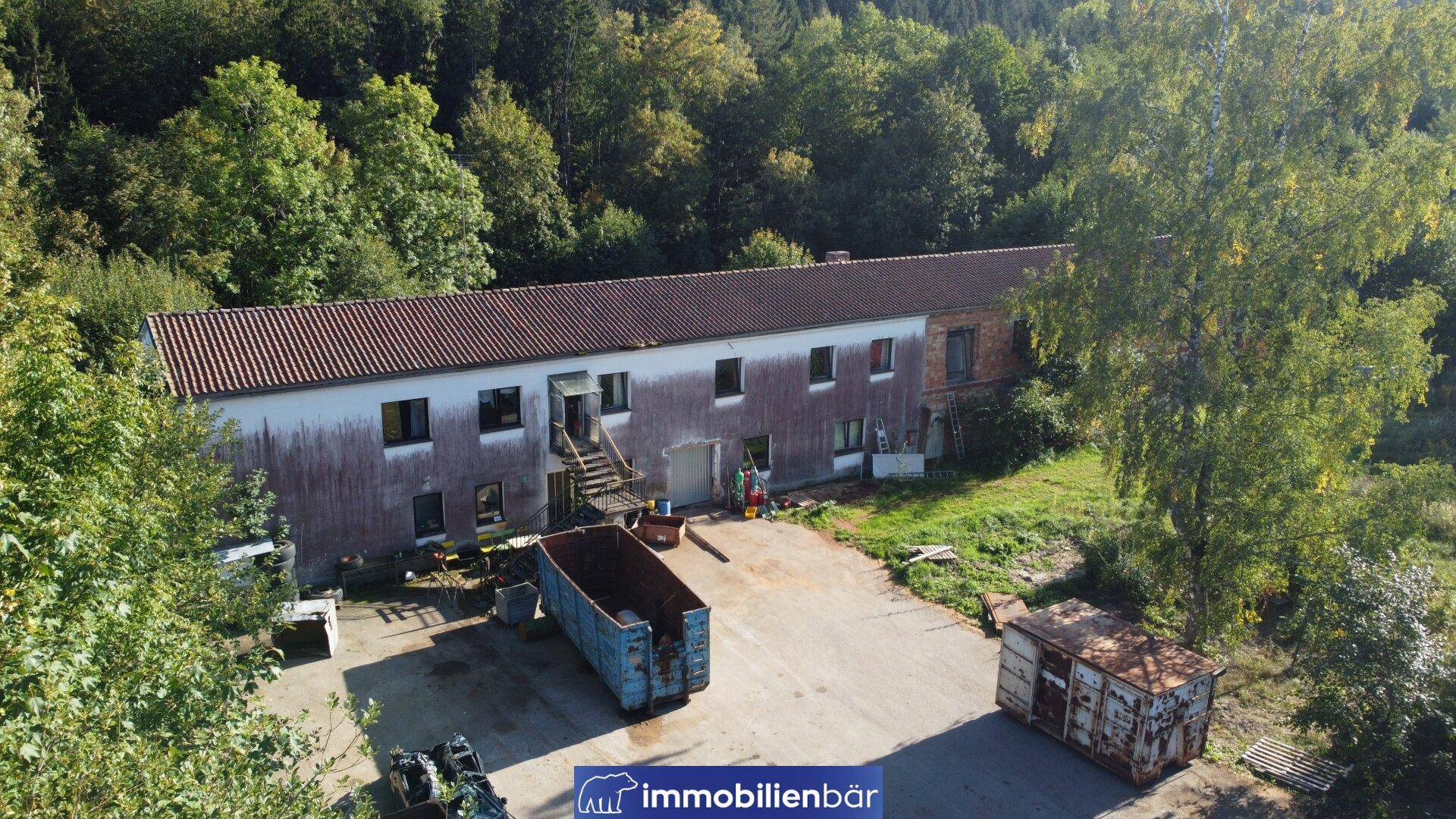 Luftaufnahme eines langen Gebäudes mit heller Fassade und Ziegeldach, umgeben von Grünflächen.