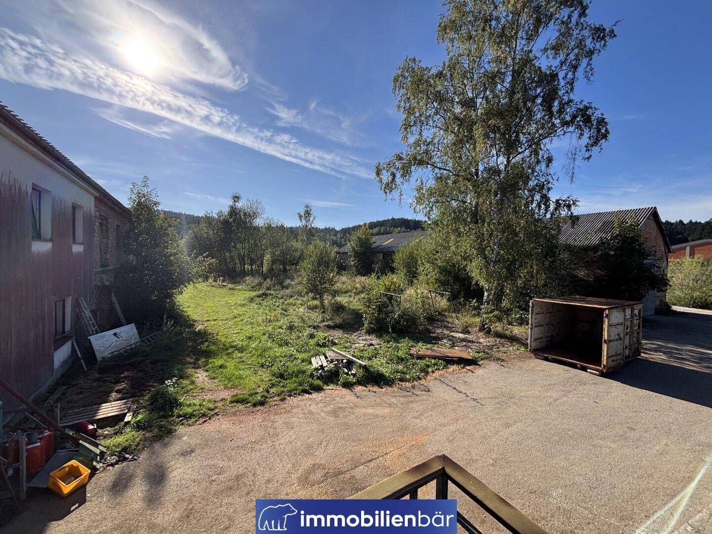Außenbereich eines Gebäudes mit sonnigem Himmel, umgeben von ungenutztem Land und Bäumen.