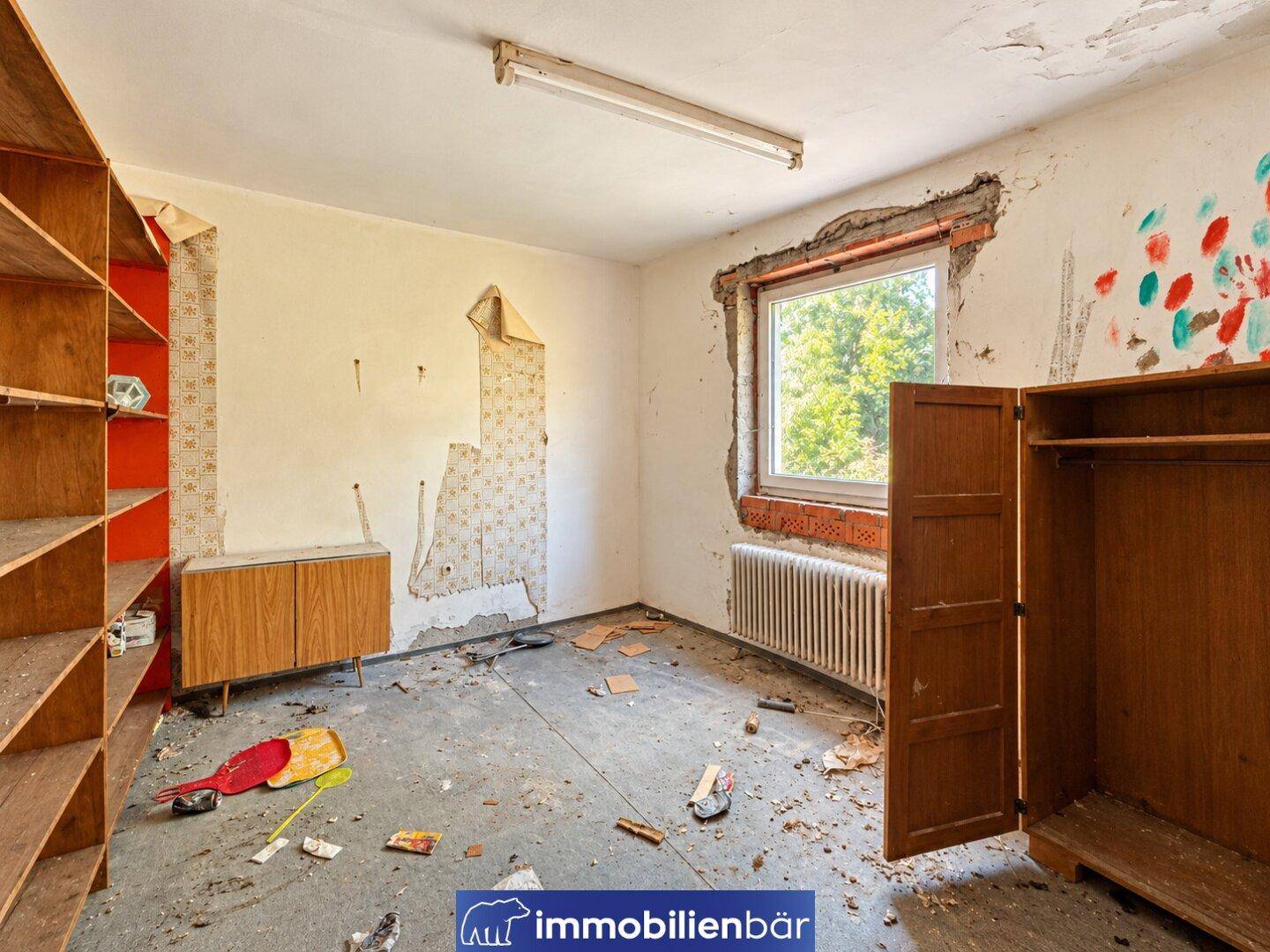 Zimmer in schlechtem Zustand mit abblätternder Tapete, freiliegenden Ziegeln und Betonboden.