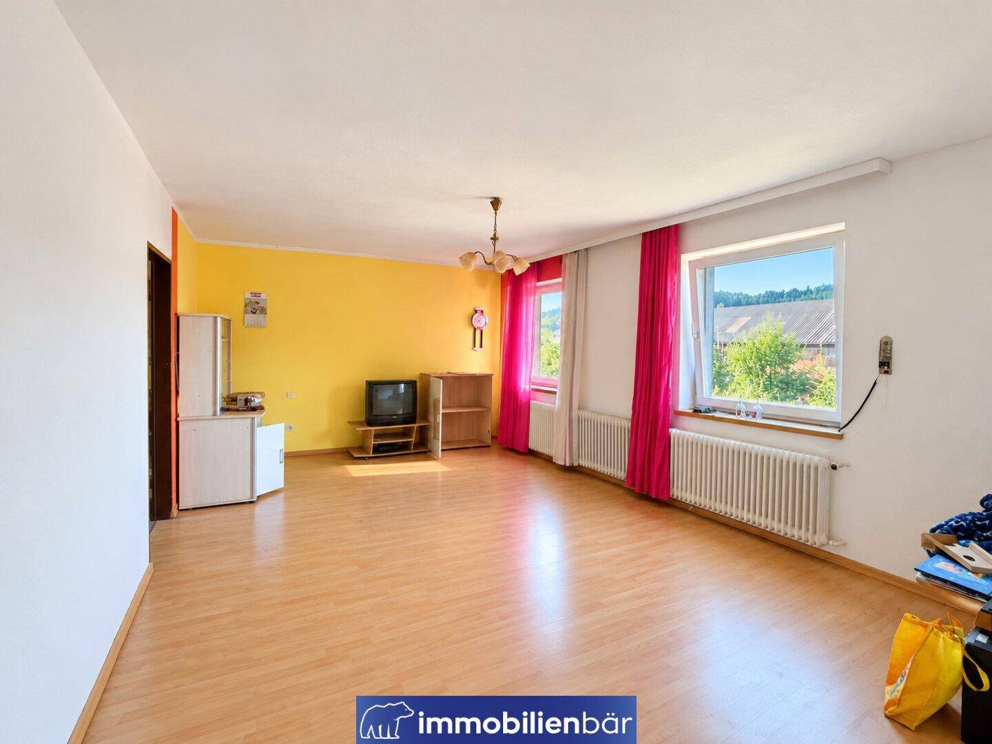Helles Wohnzimmer mit Laminatboden, gelben Wänden und zwei Fenstern mit Blick ins Grüne.