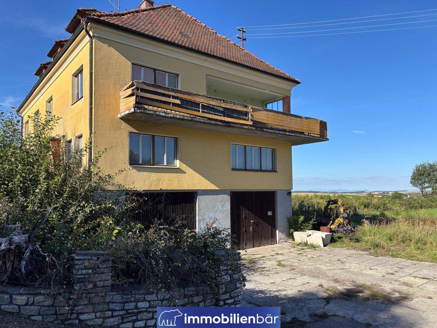 Rückansicht eines gelben Gebäudes mit einem baufälligen Balkon und Garagentoren im Erdgeschoss.