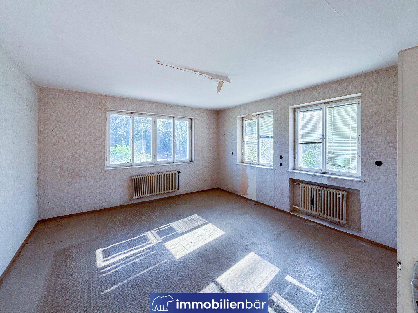 Ein leerer Raum mit abgenutztem Bodenbelag, Tapetenresten und mehreren Fenstern, die Tageslicht hereinlassen.