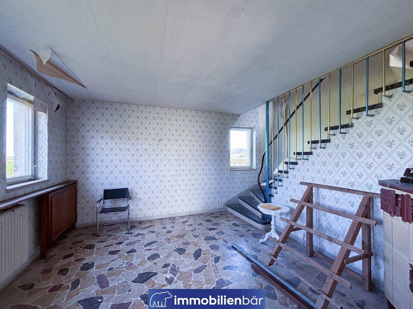 Ein Flur mit Terrazzo-ähnlichem Boden, einer geschwungenen Treppe und abgenutzten Tapeten.