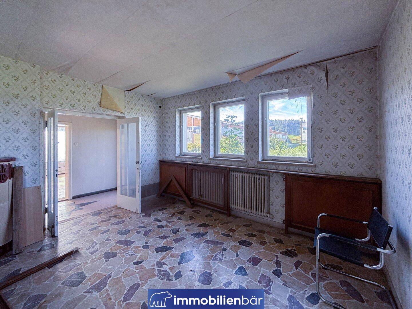 Ein Raum mit Terrazzo-Boden, mehreren Fenstern und einer Tür, die zu einem weiteren Zimmer führt.
