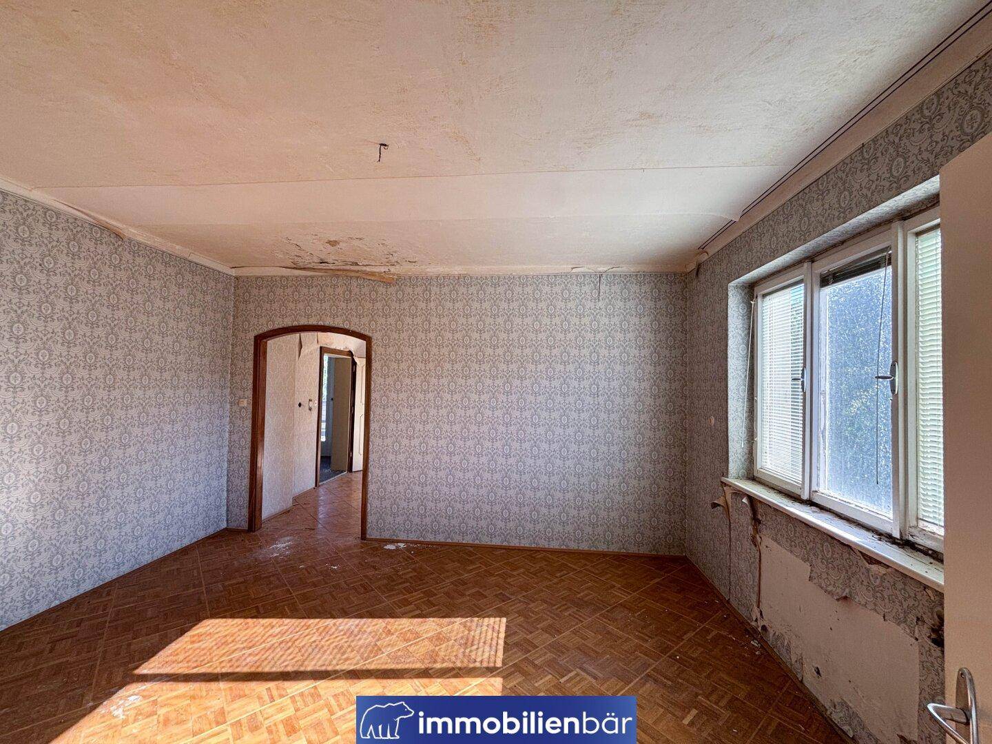 Ein leerer Raum mit abgenutztem Parkettboden, alten Tapeten und einem Fenster mit Blick nach draußen.