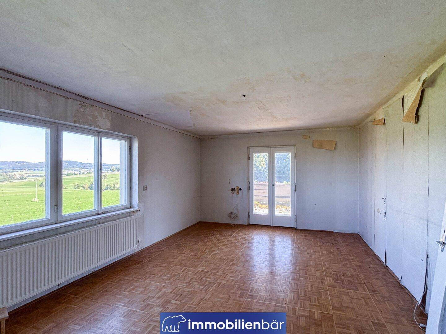 Ein heller Raum mit Parkettboden, großen Fenstern und einer Tür, die auf einen Balkon führt.