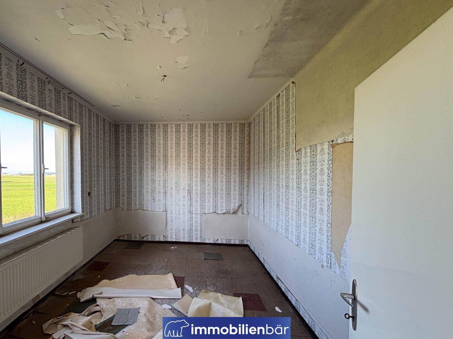 Ein Raum mit abblätternden Tapeten, altem Bodenbelag und einem Fenster mit Blick auf die Landschaft.