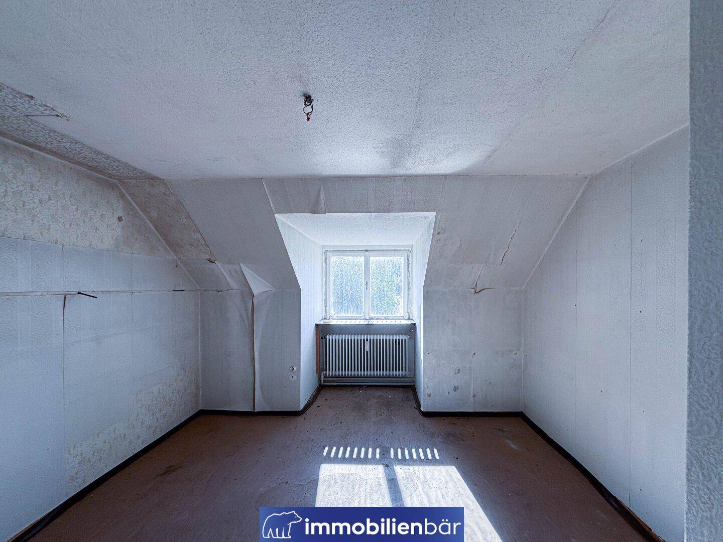 Ein Dachzimmer mit schrägen Wänden, einem Dachfenster und abgenutzten Oberflächen.