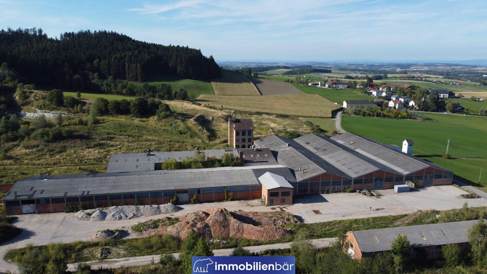Weitläufige Ansicht eines Industriegebäudekomplexes in ländlicher Umgebung mit Feldern und einem Wald im Hintergrund.