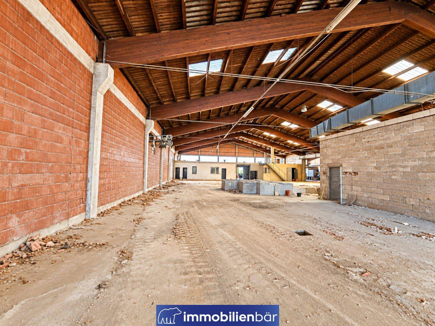 Lange Industriehalle mit roten Ziegelwänden, Holzdachkonstruktion und einem unbefestigten Boden, der viel Platz bietet.