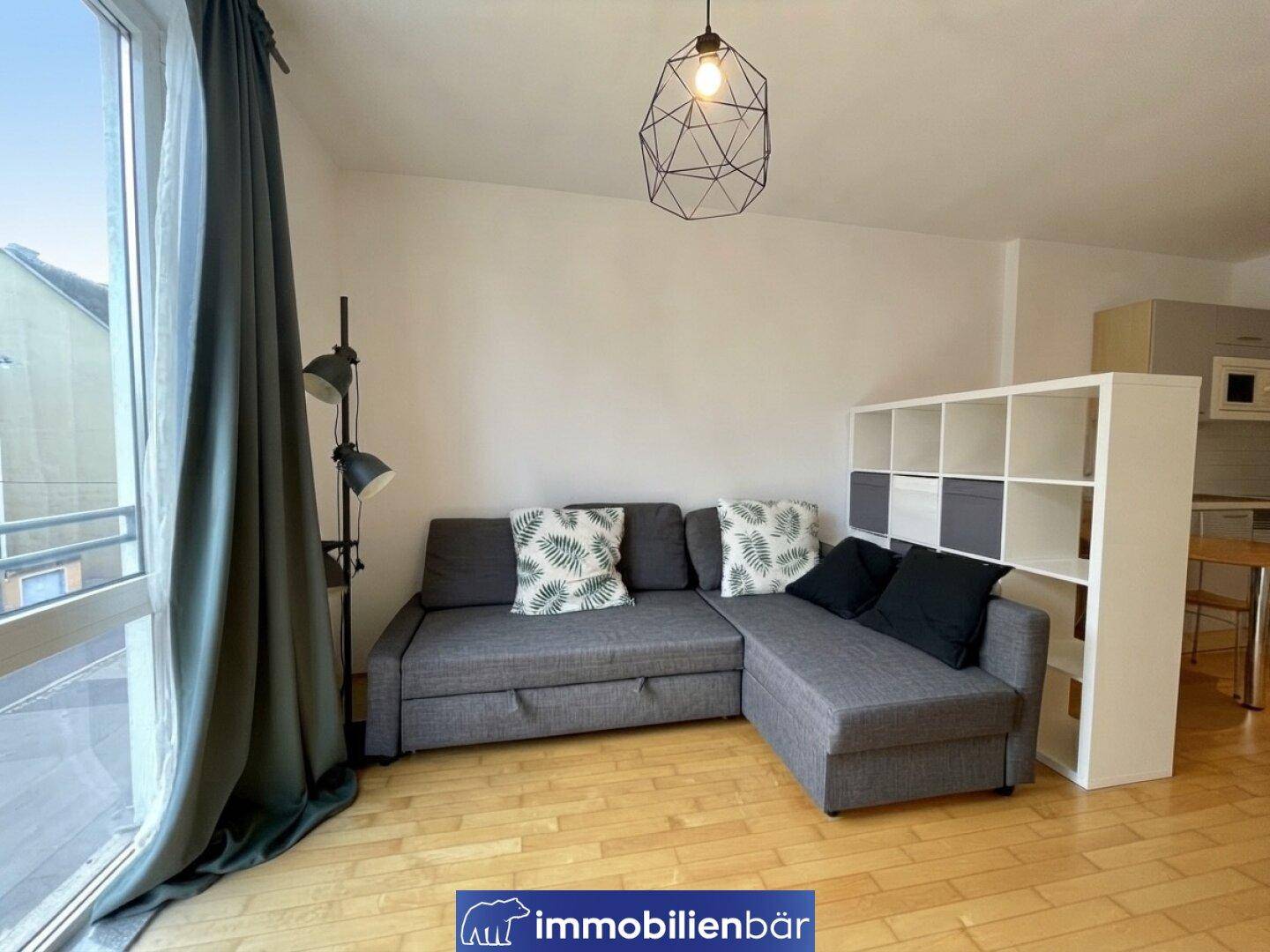 Wohnbereich mit Ecksofa, Regal und Zugang zu einem Balkon durch eine große Glastür.