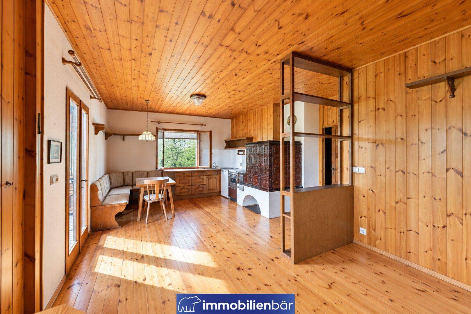 Gemütlicher Wohn-Essbereich mit Holzboden und -decke, inklusive traditionellem Kachelofen und Küchenzeile.