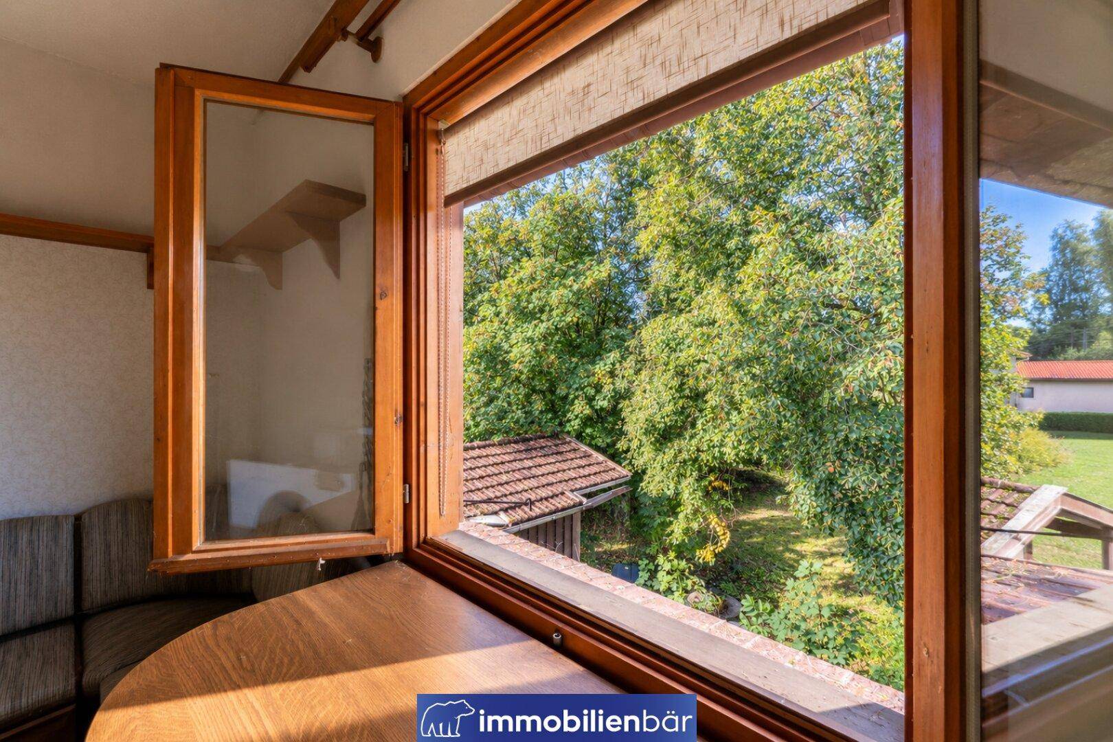 Fenster mit Holzrahmen bietet einen grünen Ausblick auf Bäume und ein kleines Nebengebäude.