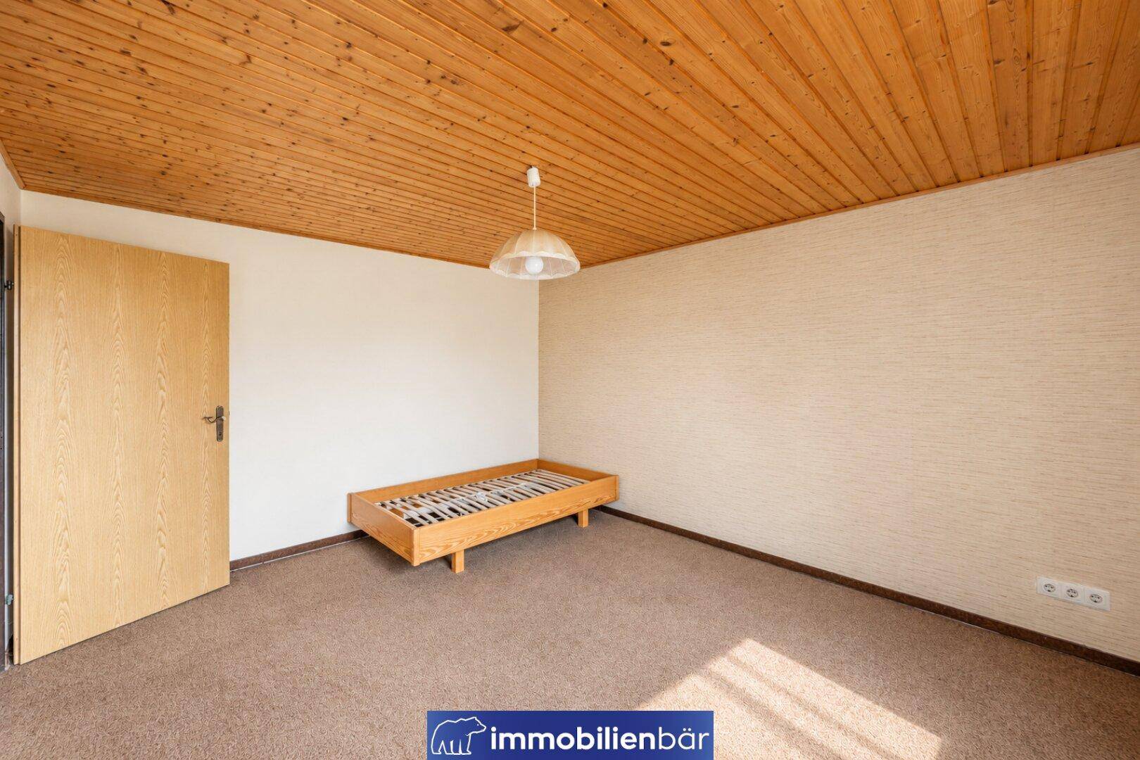 Leeres Schlafzimmer mit Teppichboden, Holzdecke und einem einfachen Bettgestell.