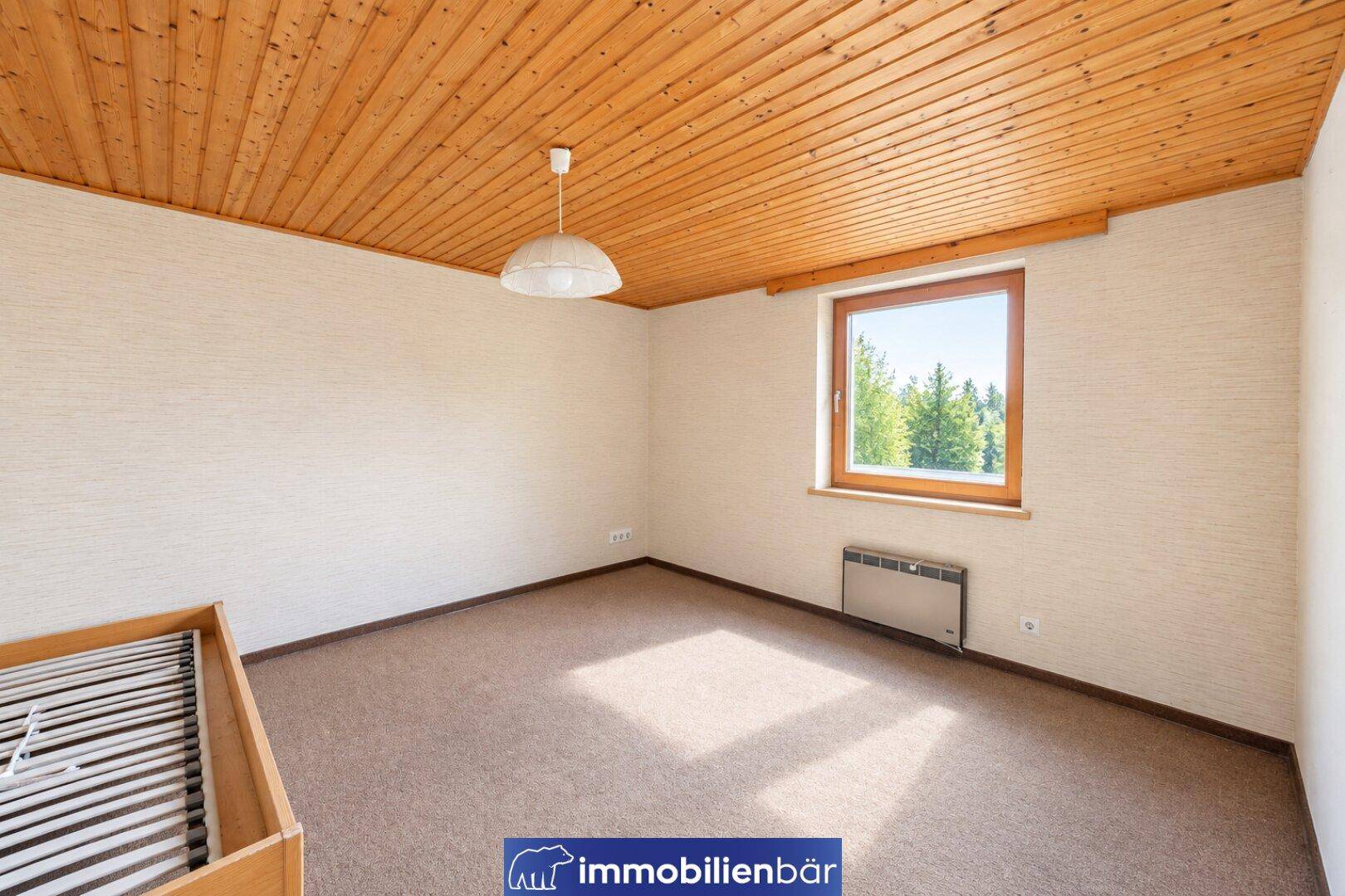 Helles Schlafzimmer mit Teppichboden, Holzdecke und einem Fenster mit Blick ins Grüne.