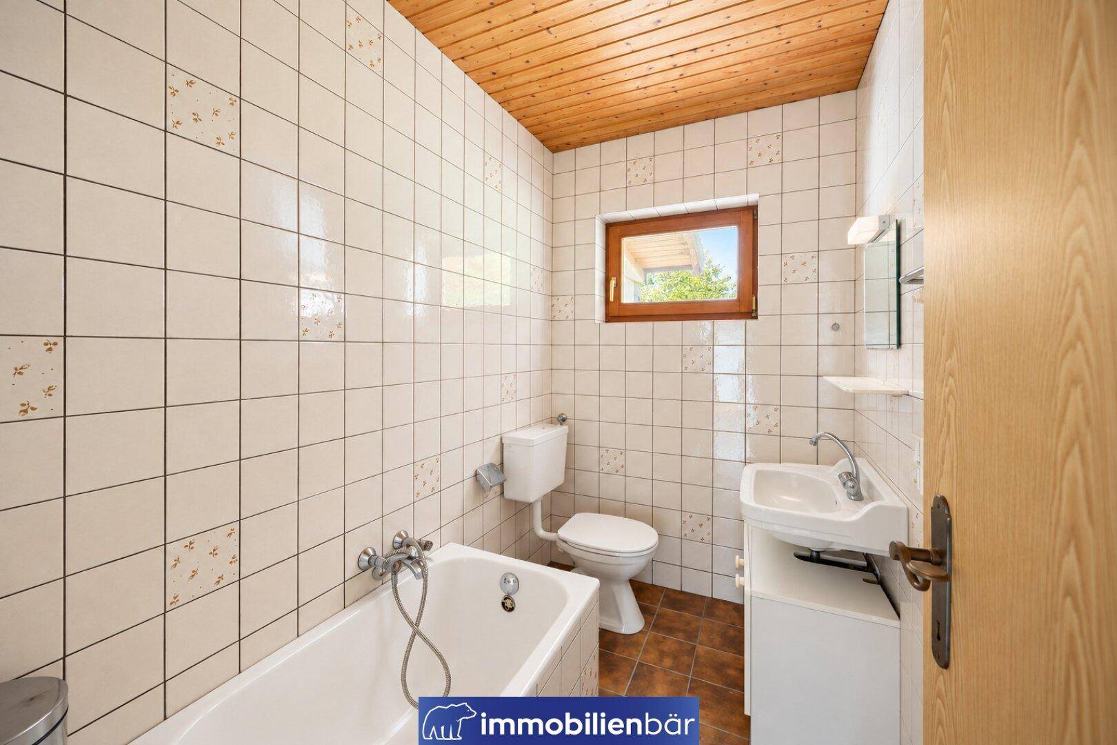 Badezimmer mit weißen Fliesen, Badewanne, Toilette und Waschbecken, sowie einem Fenster.