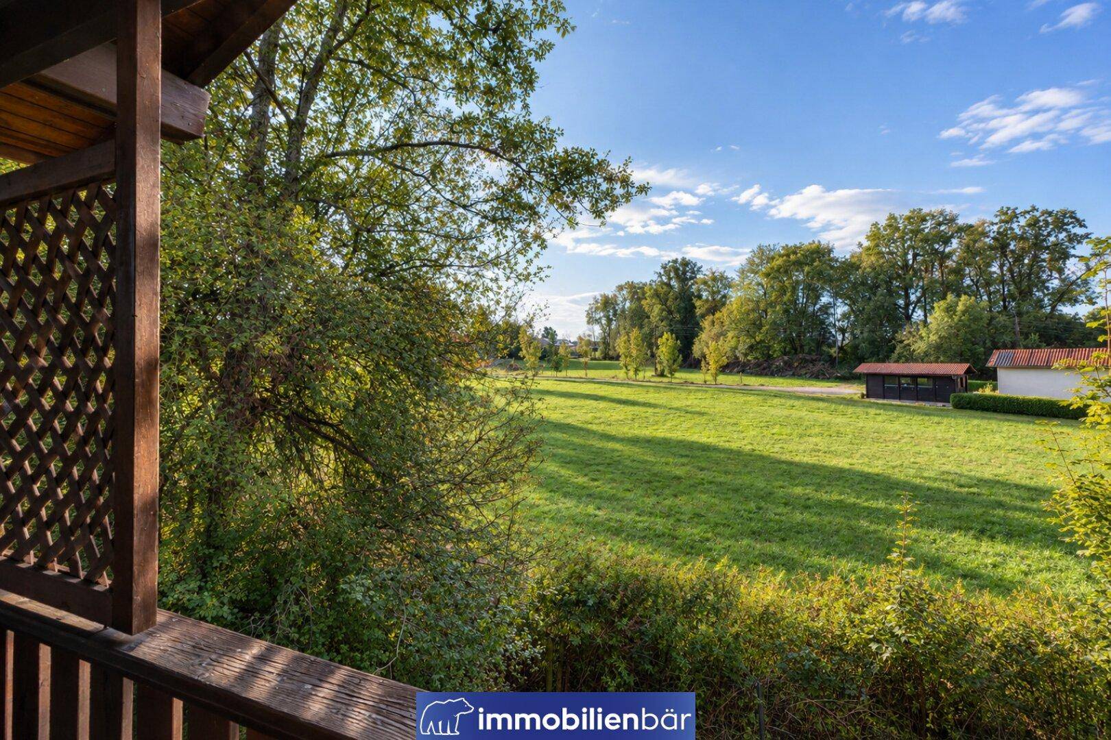 Weitläufiger Blick von einem Balkon auf eine große grüne Wiese und entfernte Bäume.