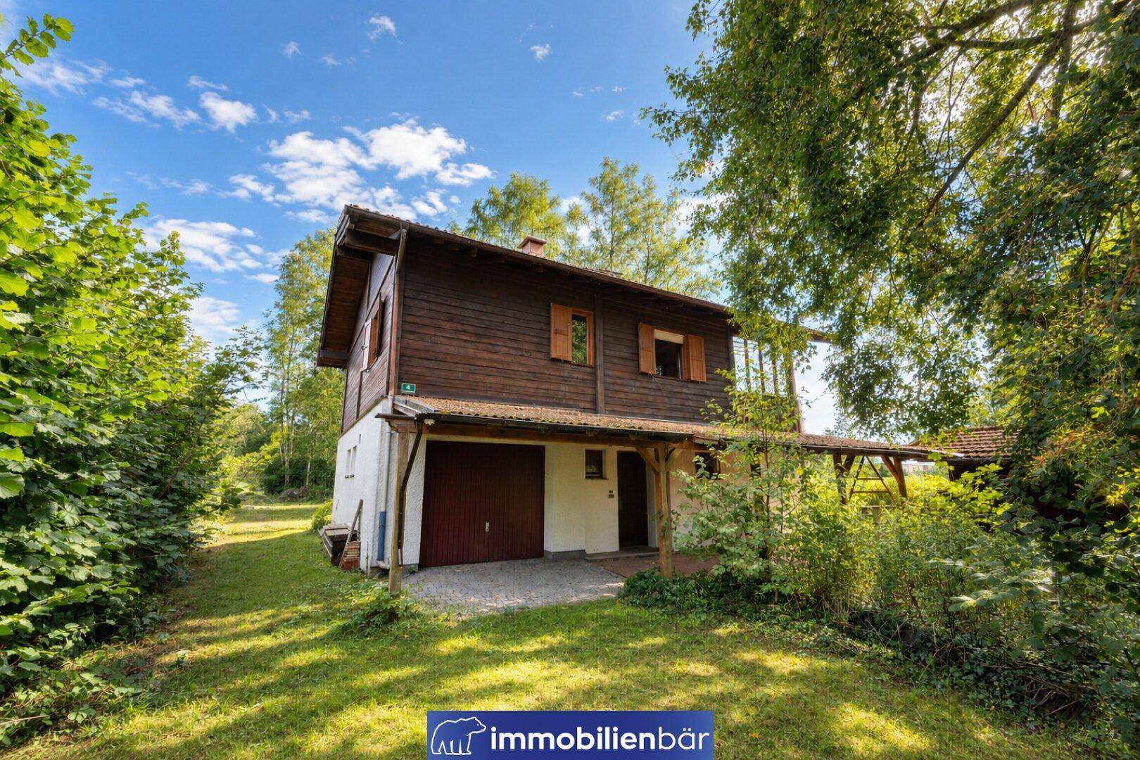 Rustikales Holzhaus mit integrierter Garage und umgebendem Gartenbereich unter hellem Himmel.
