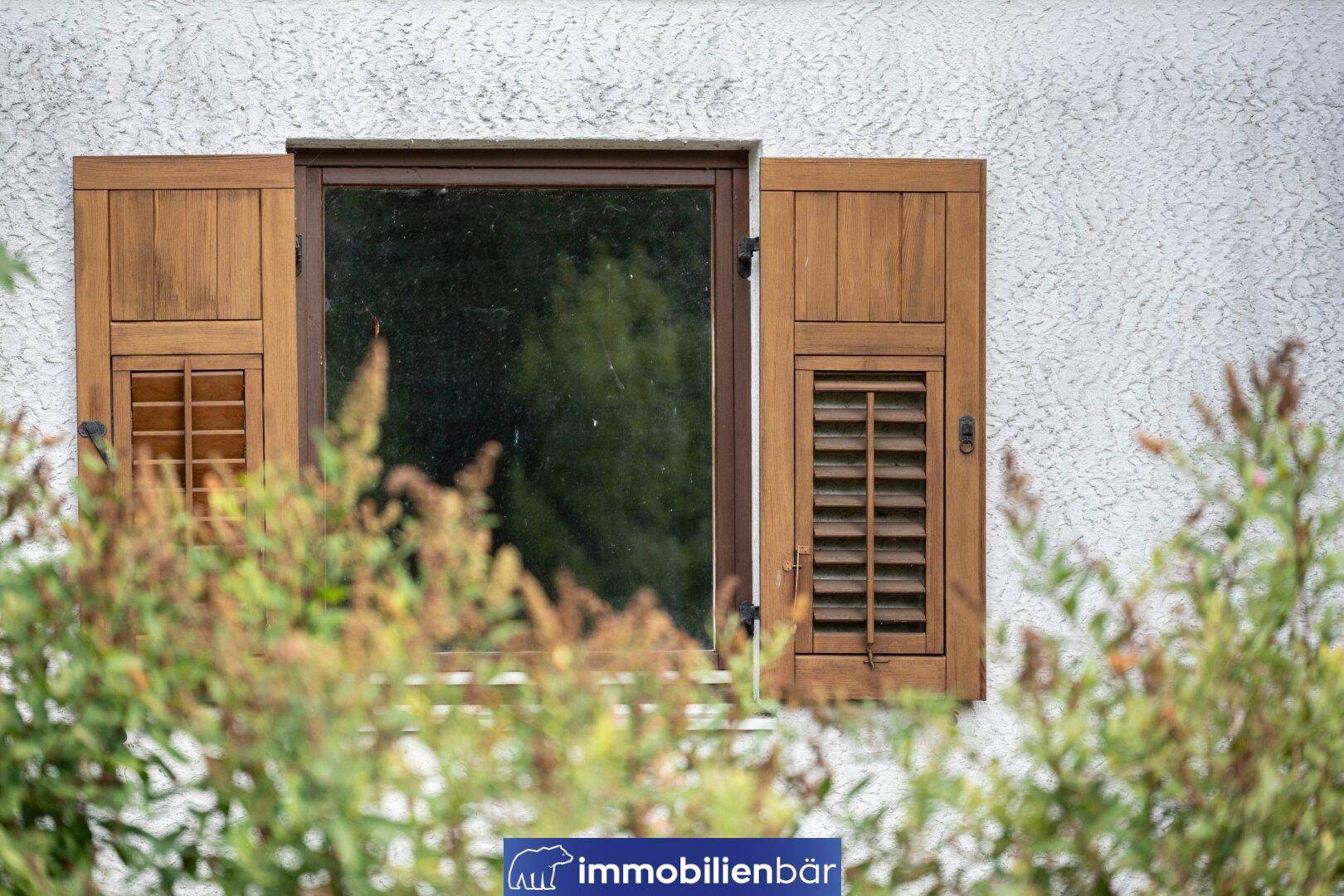 Fenster mit geöffneten Holzläden an einer weiß verputzten Fassade, umgeben von Grün.
