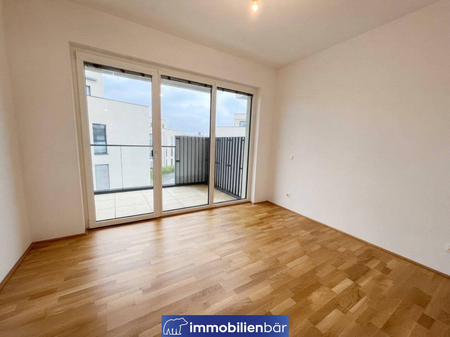 Leeres Zimmer mit Parkettboden und breiter Glasfront, die viel Tageslicht hereinlässt.