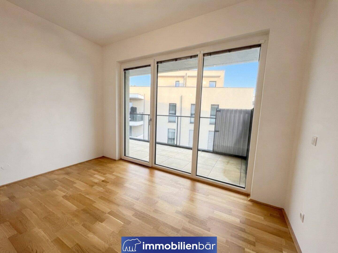 Heller Raum mit Holzboden und großen Fenstern, die einen Blick auf andere Gebäude bieten.