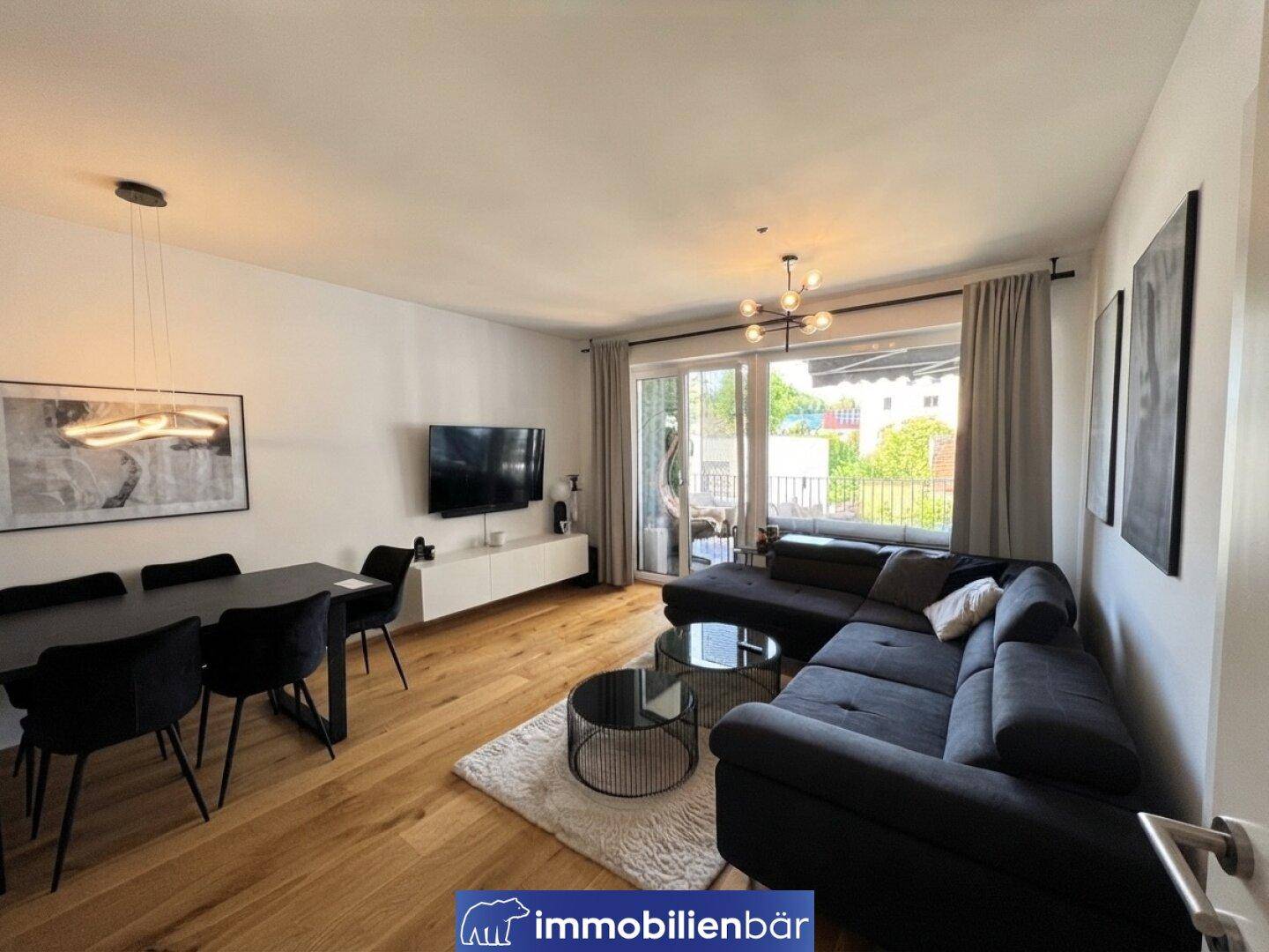 Moderner Wohn-Essbereich mit dunklem Sofa, Holzboden und Zugang zum Balkon.