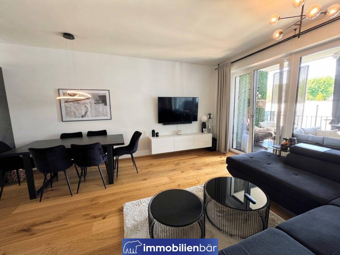 Heller Wohn-Essbereich mit dunklem Sofa, Holzboden und großem Fenster zum Balkon.