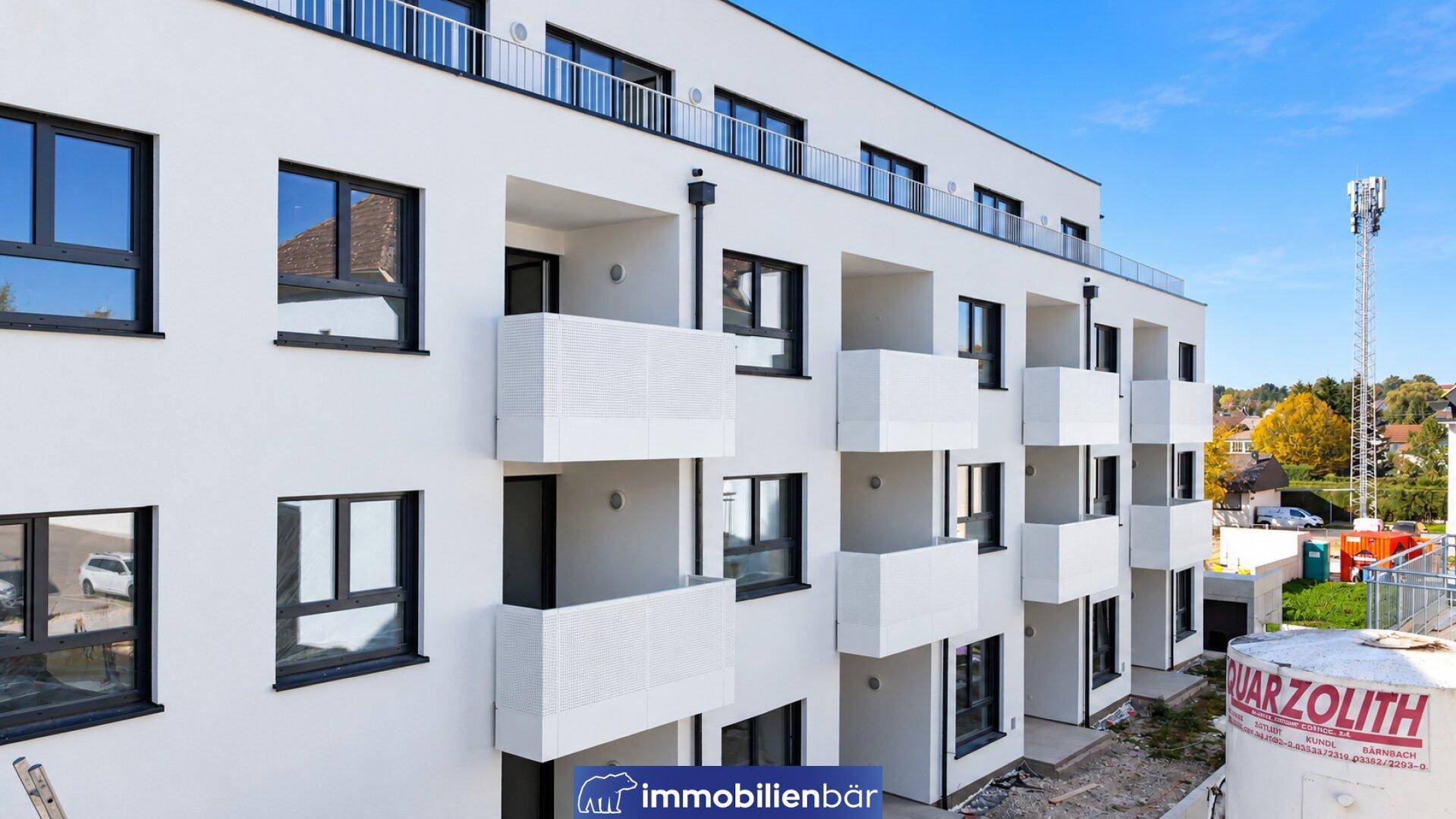 Moderne weiße Fassade eines Neubaus mit zahlreichen Balkonen unter blauem Himmel.