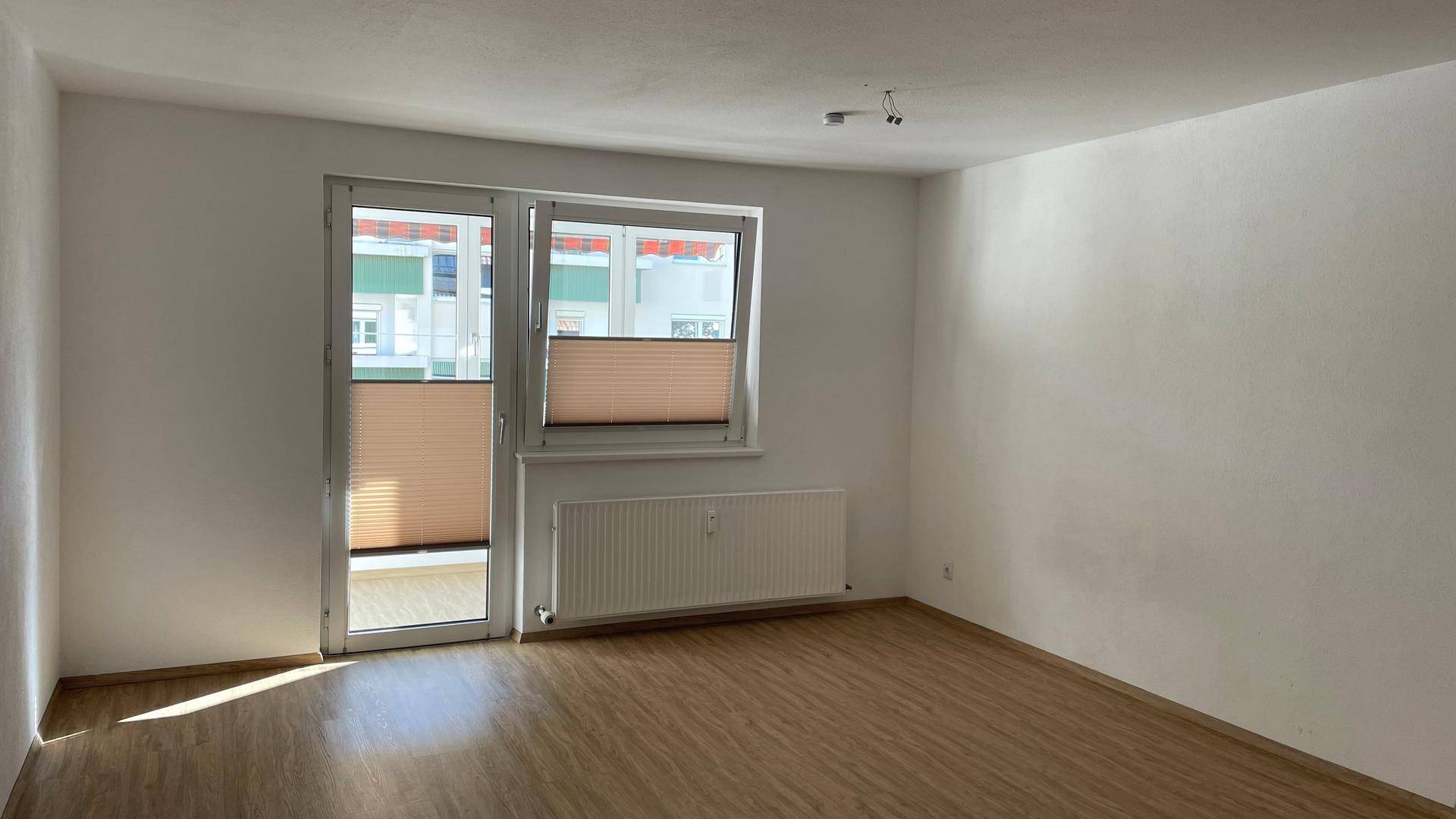 Geräumiger, leerer Raum mit Laminatboden und großem Fenster mit Heizkörper.