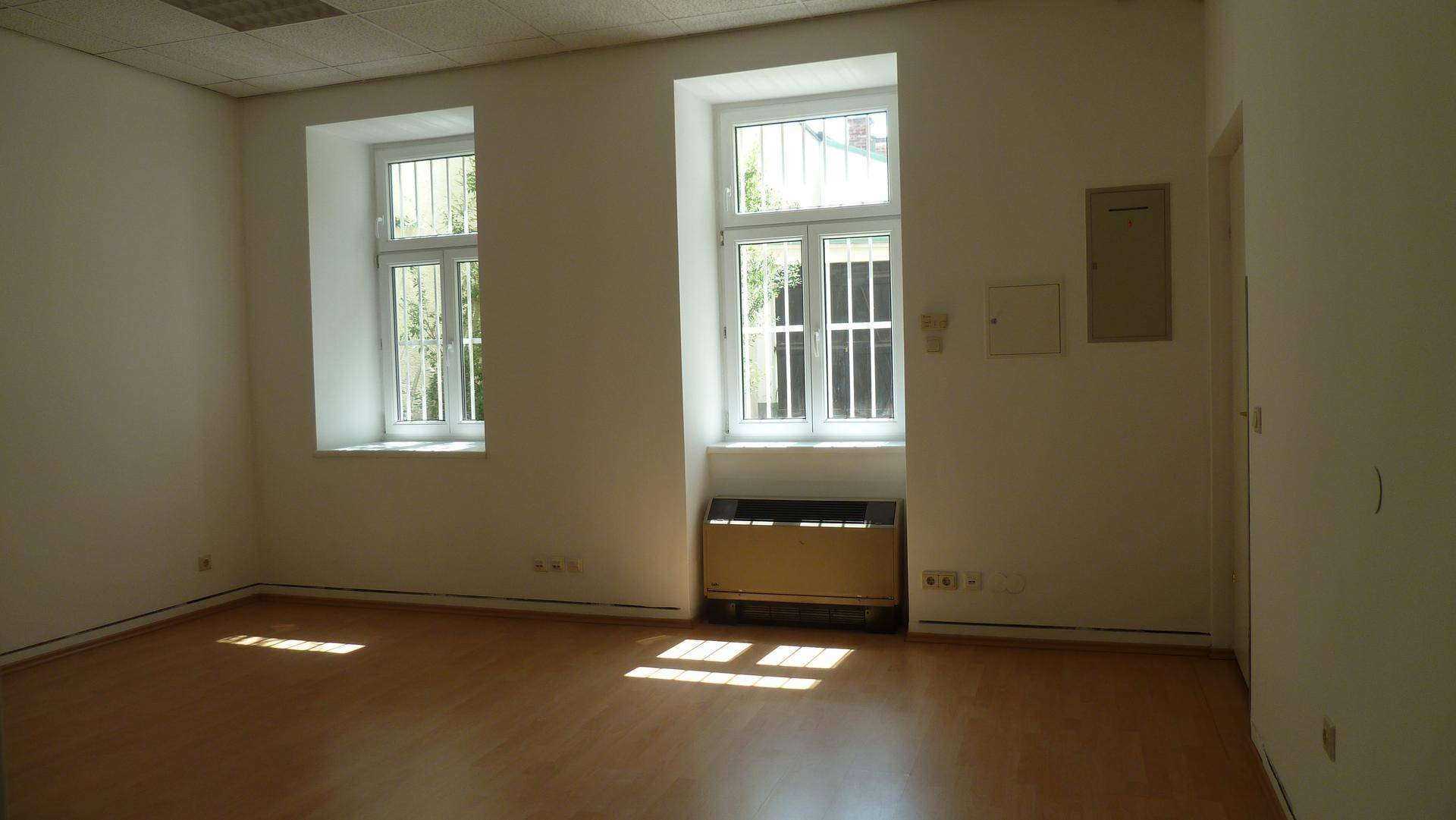 Heller Büroraum mit Holzfußboden, zwei Fenstern mit Gittern und einer Heizung unter dem Fenster.