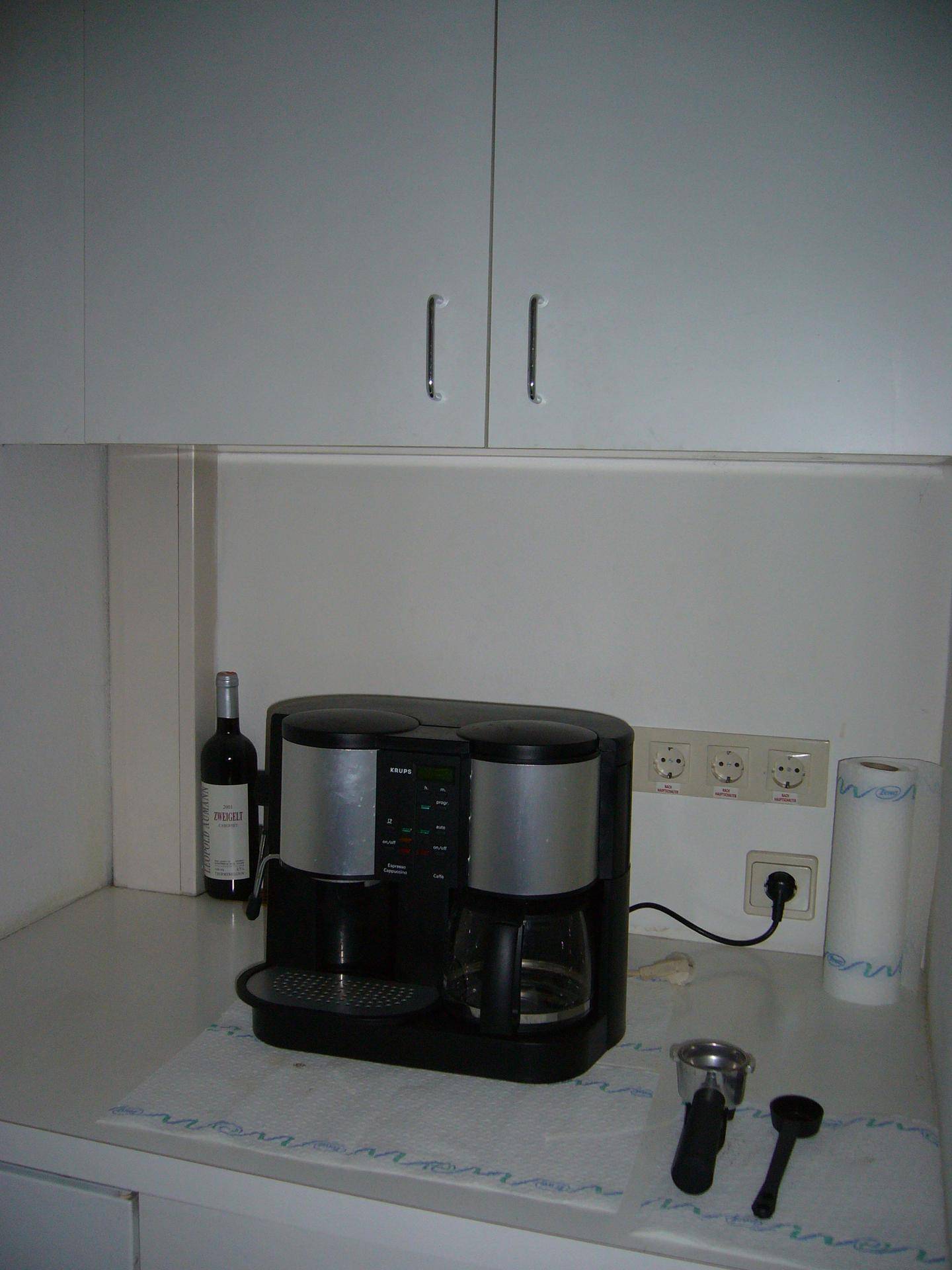 Kleine Küchenzeile mit Kaffeemaschine, weißen Schränken und Steckdosen an der Wand.
