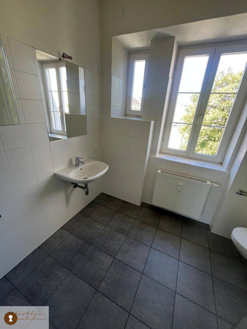 Helles Badezimmer mit Fenster, Waschbecken und dunklen Bodenfliesen.