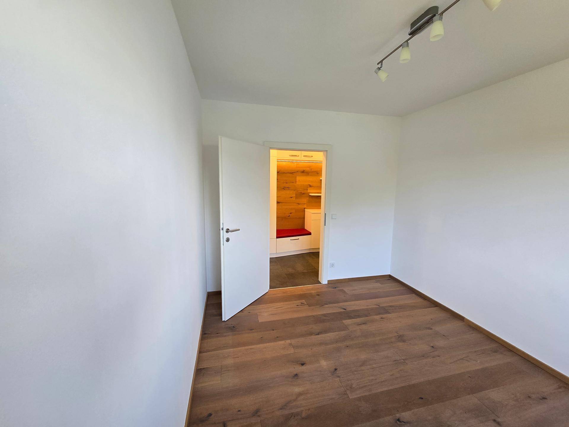 Leeres Zimmer mit Holzboden und Zugang zum Flur mit integrierter Garderobe.