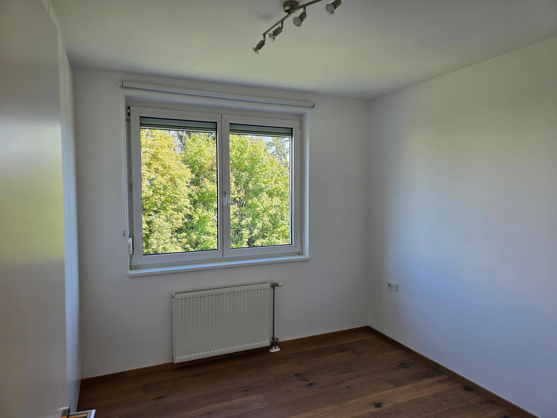 Geräumiges Zimmer mit Holzboden und Fenster mit Blick auf die Baumwipfel.