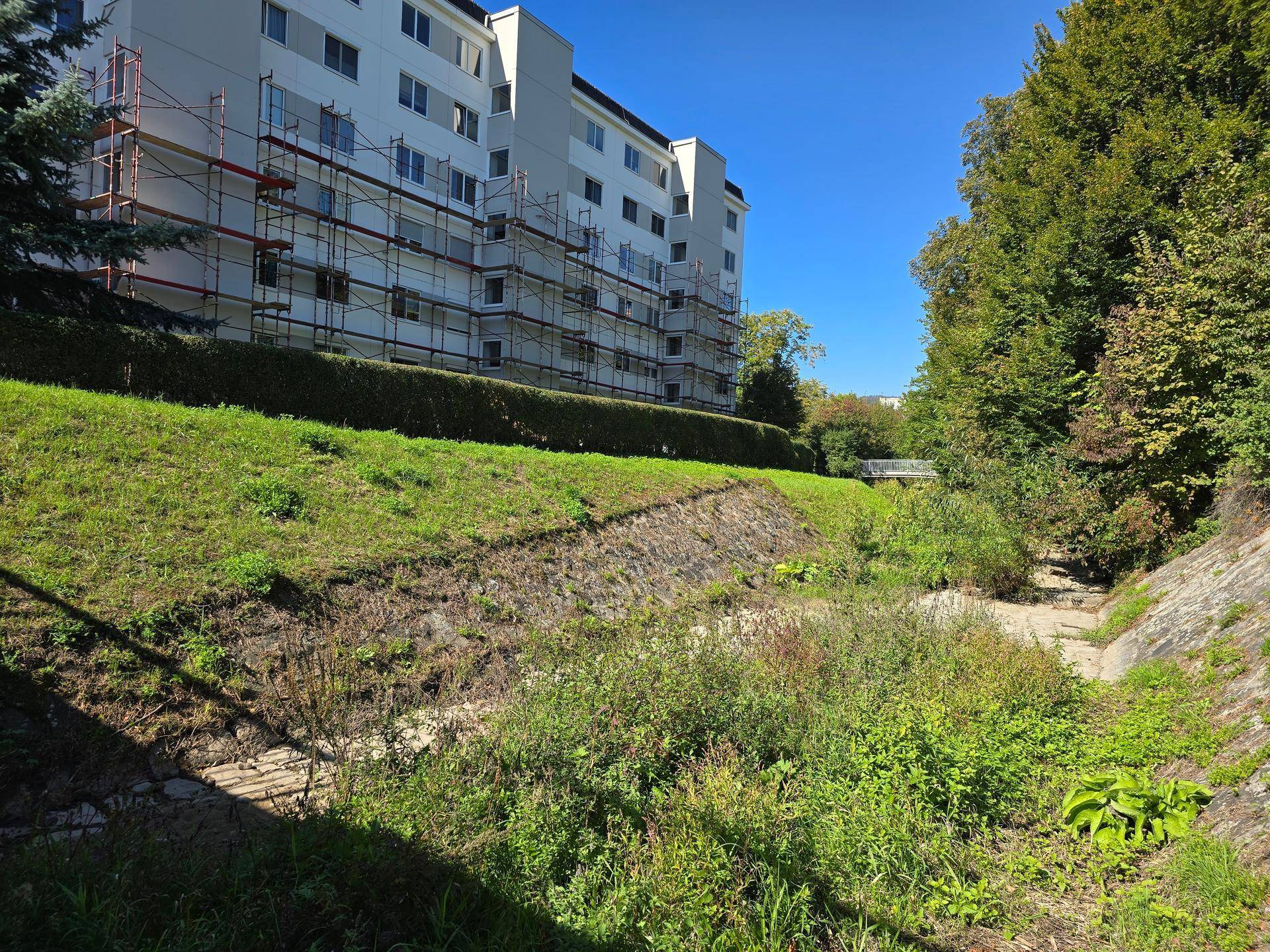 Wohngebäude mit Gerüst an der Fassade, angrenzend an eine natürliche, bewachsene Böschung.