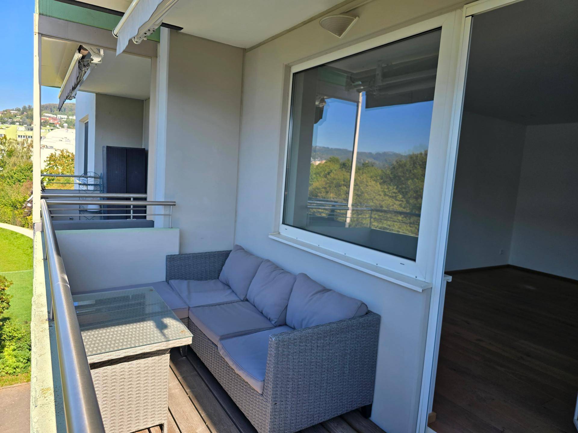 Möblierter Balkon mit Rattan-Sofa und Holzboden, angrenzend an einen leeren Innenraum.