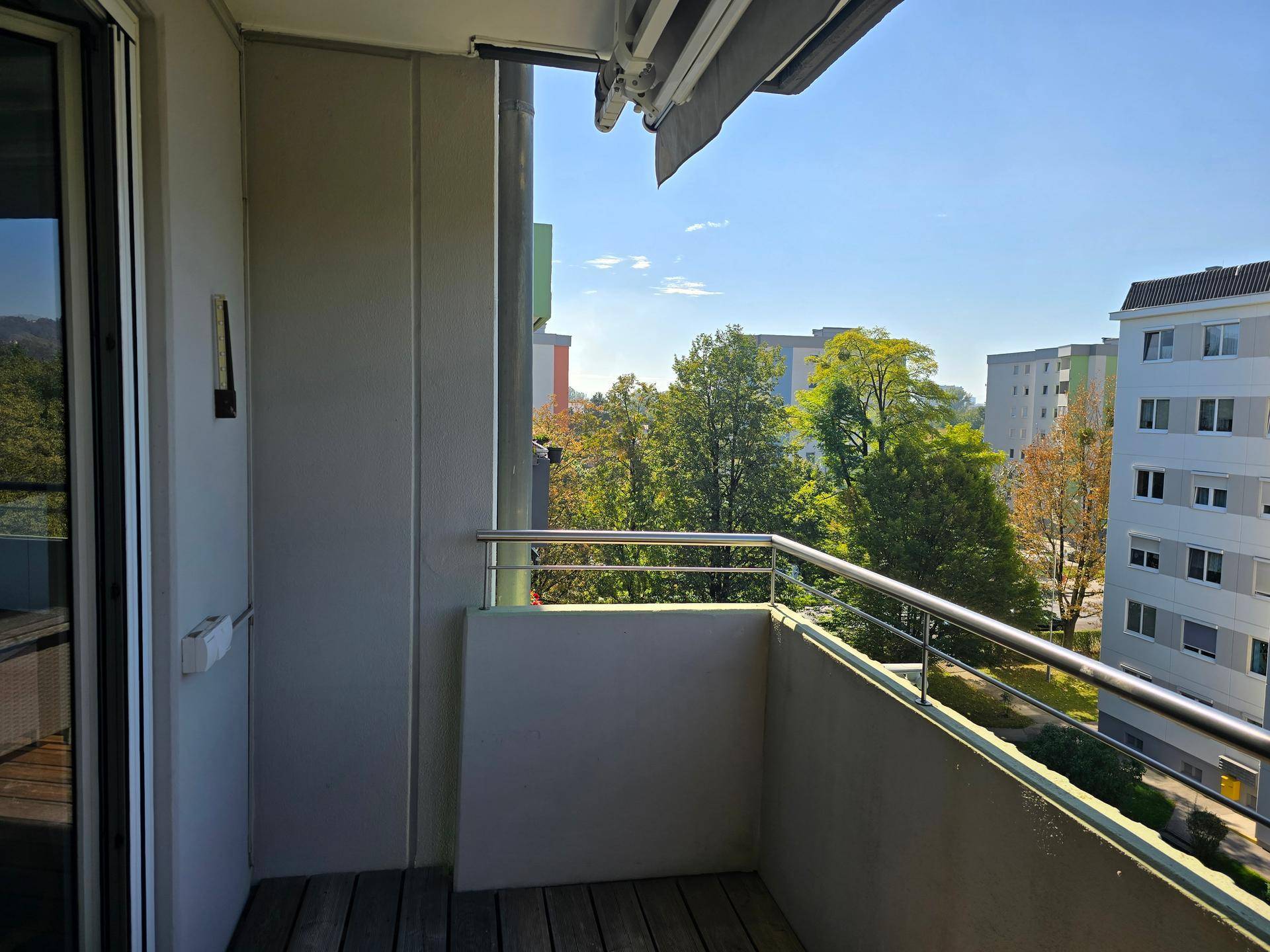 Leerer Balkon mit Metallgeländer und Blick auf die umliegenden Gebäude und grüne Bäume.