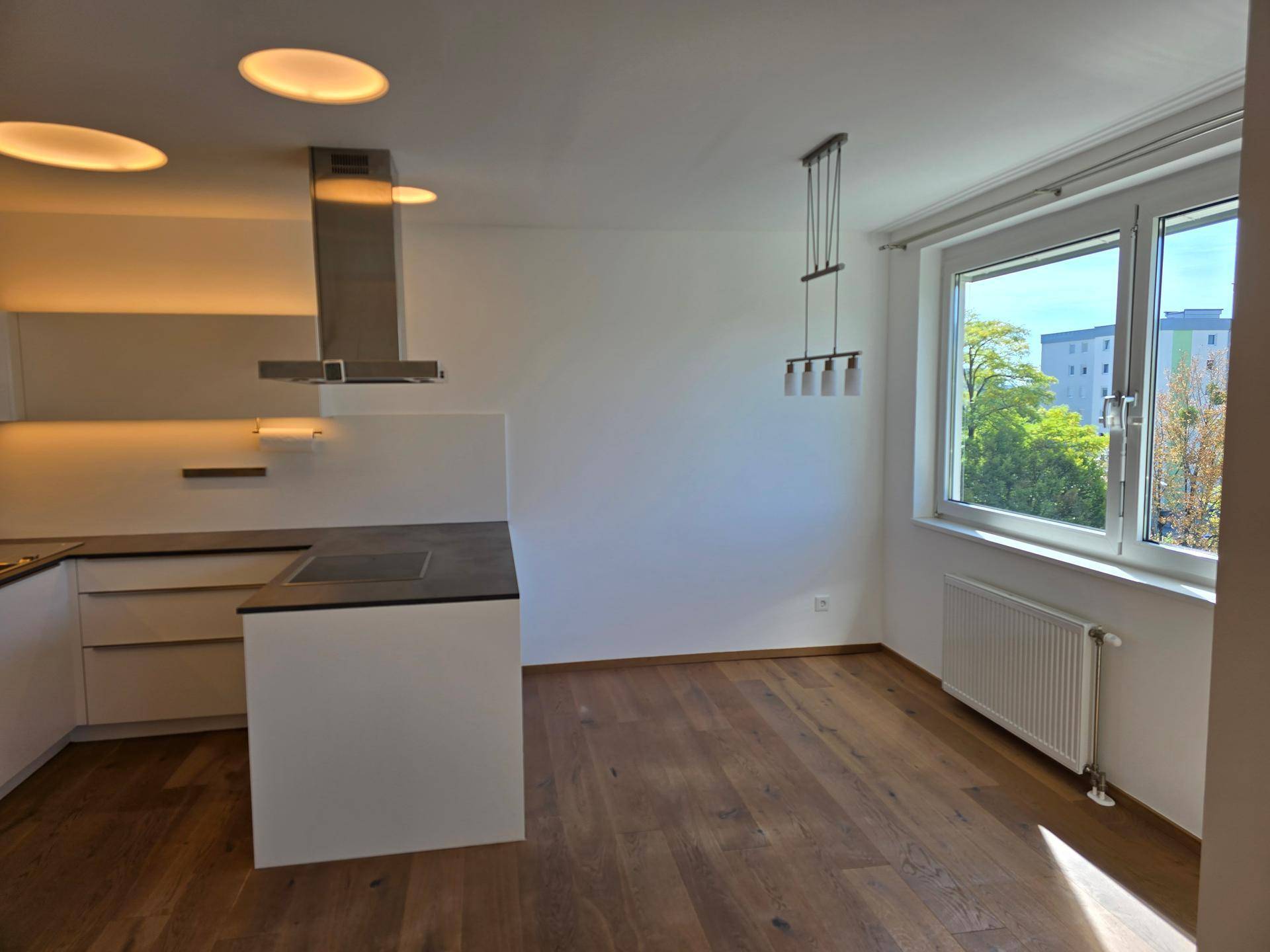 Heller Wohnbereich mit Holzboden, großem Fenster und moderner Deckenbeleuchtung.