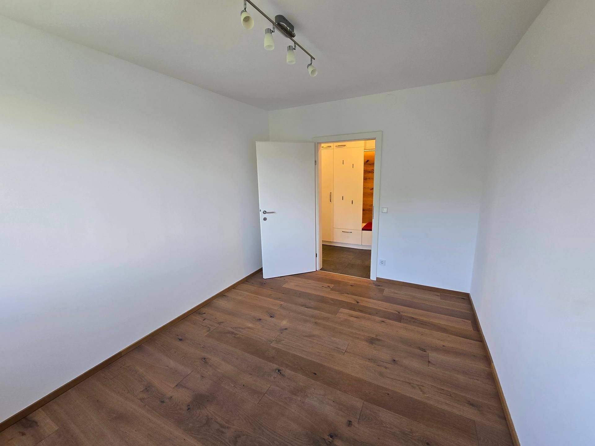 Helles Zimmer mit Holzboden und offener Tür zum Flur mit Einbauschränken.