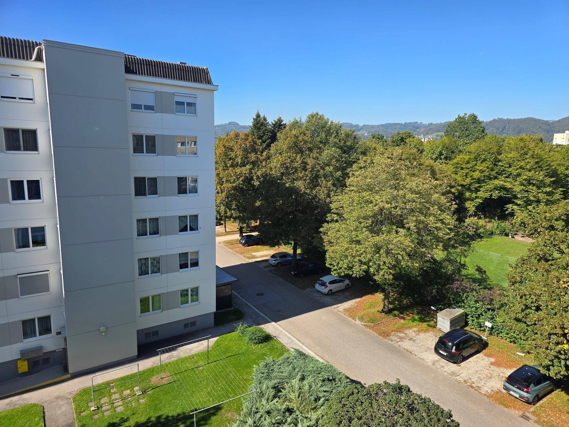 Blick auf ein Wohngebäude mit Parkplätzen und einer grünen Umgebung unter blauem Himmel.