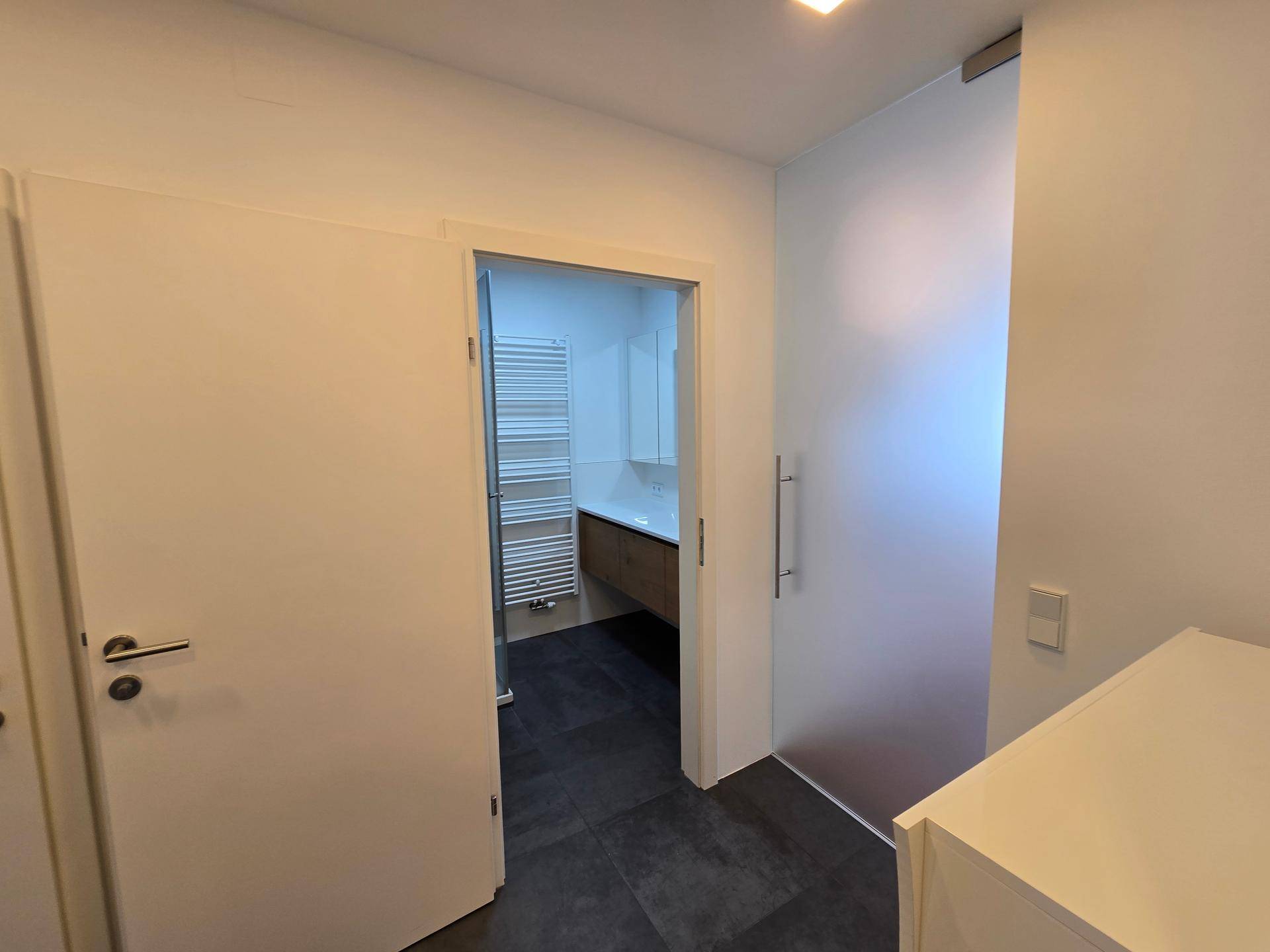 Flur mit dunklen Fliesen, Blick in ein modernes Badezimmer und eine Milchglastür.