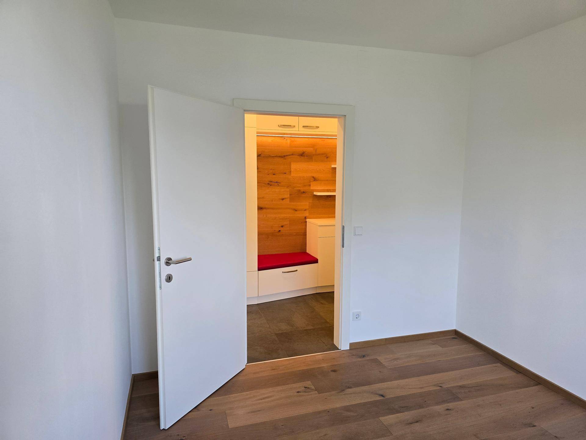 Helles Zimmer mit Holzboden und offener Tür, die den Blick auf die Garderobe freigibt.