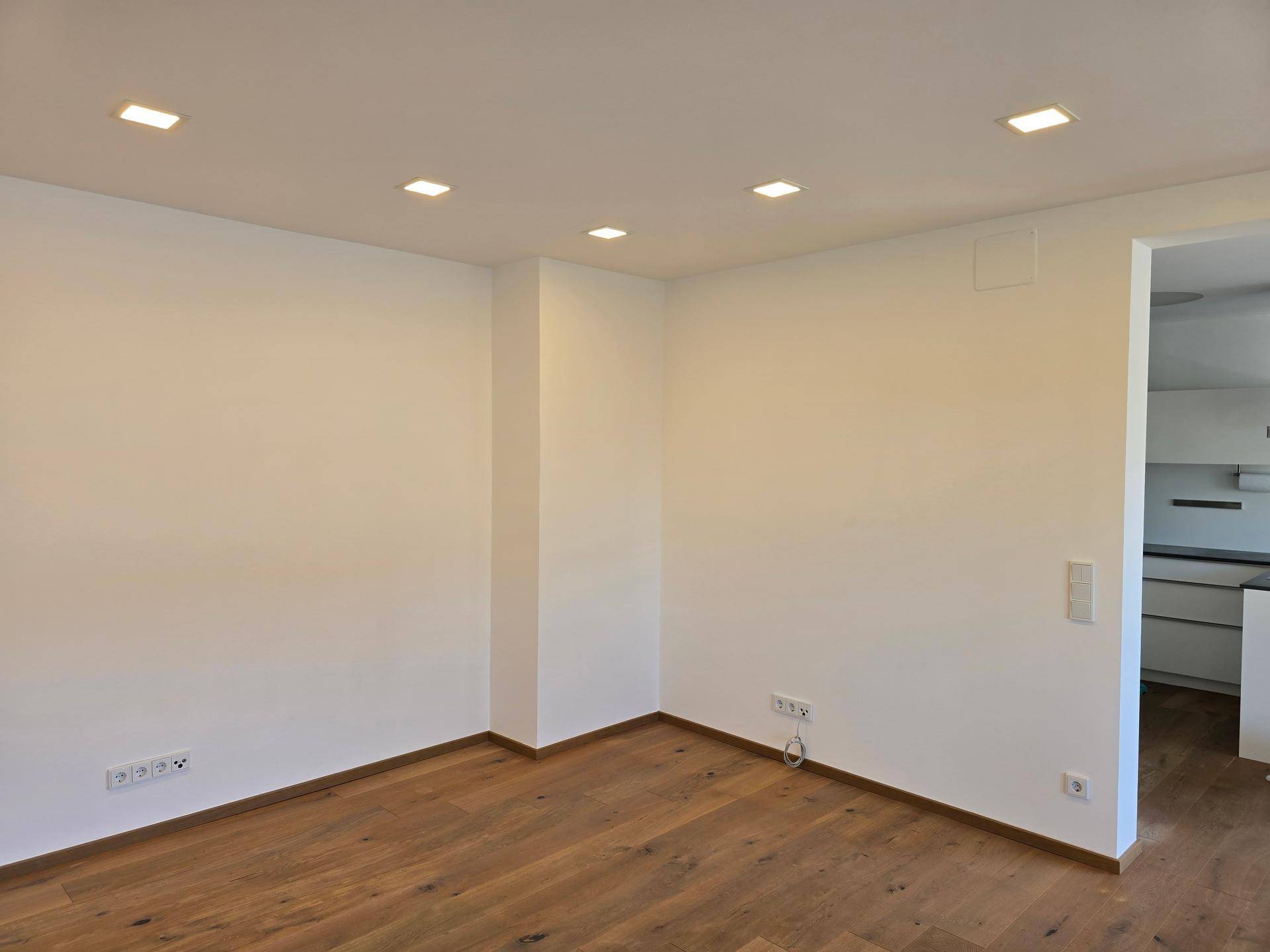 Heller Wohnraum mit Holzboden und moderner Deckenbeleuchtung, Blick in den Küchenbereich.