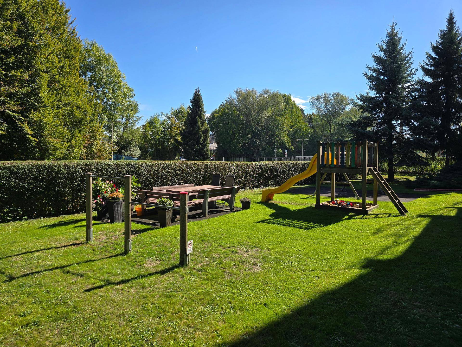 Gemeinschaftlicher Spielplatz mit Rutsche, Picknicktisch und viel Grünfläche unter blauem Himmel.
