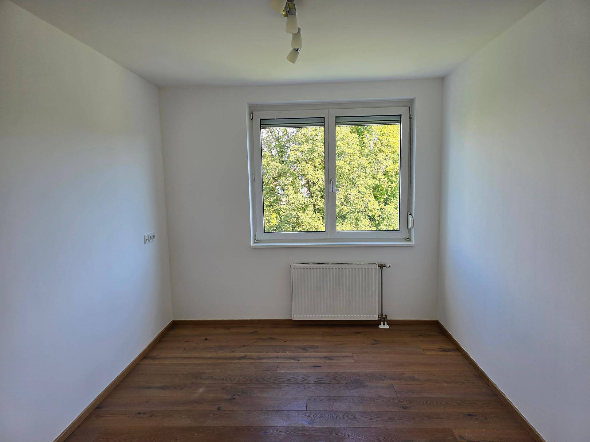 Helles Zimmer mit Holzboden und Blick ins Grüne durch das Fenster.