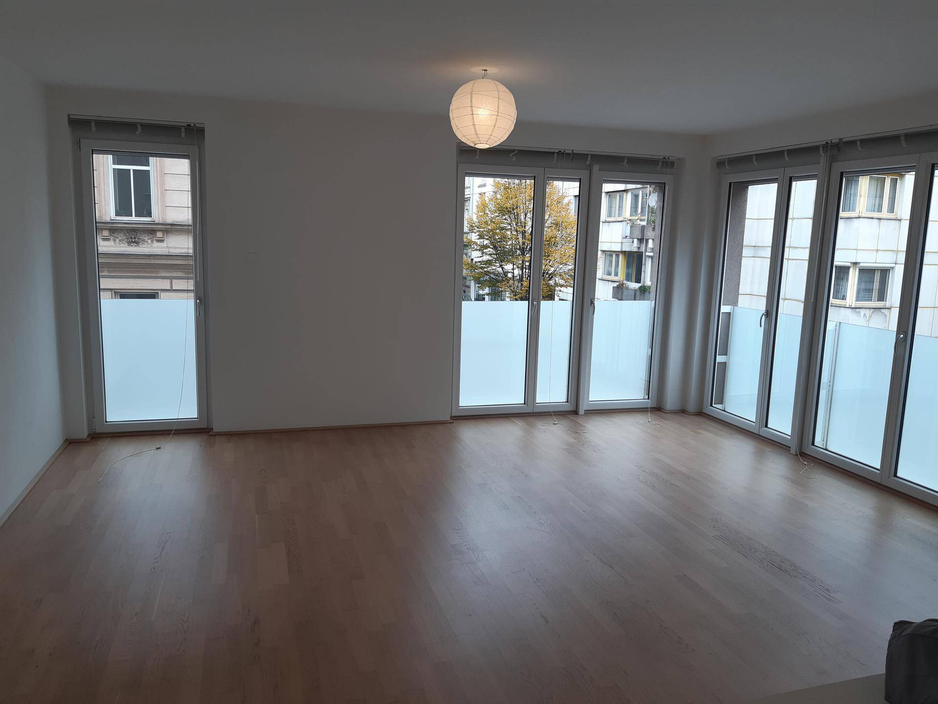 Heller Wohnbereich mit großen Fenstern, Holzboden und Blick auf die umliegenden Gebäude und Bäume.